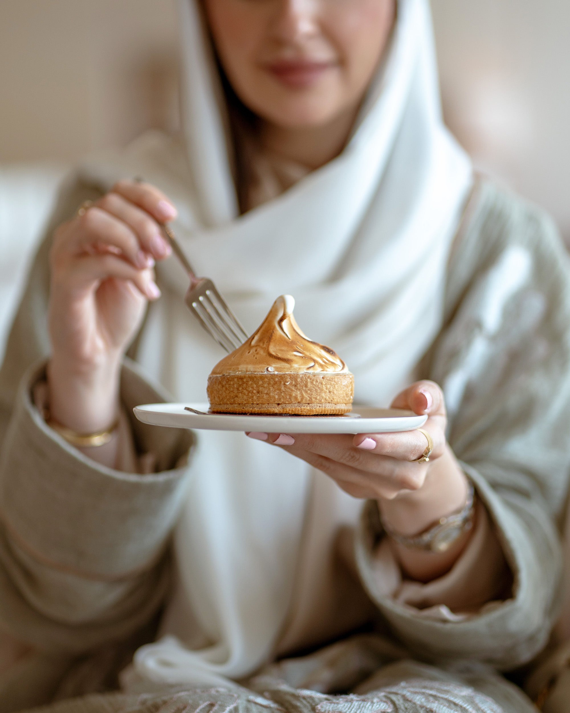 Person holding a dessert with a fork, wearing a white hijab and beige outfit.