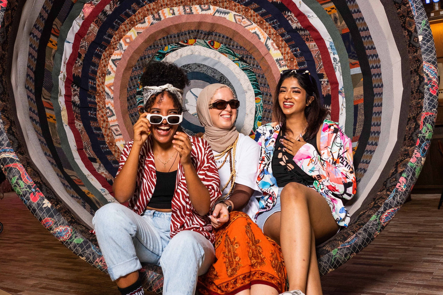 Three women sitting on a large, colorful circular chair in an indoor setting.